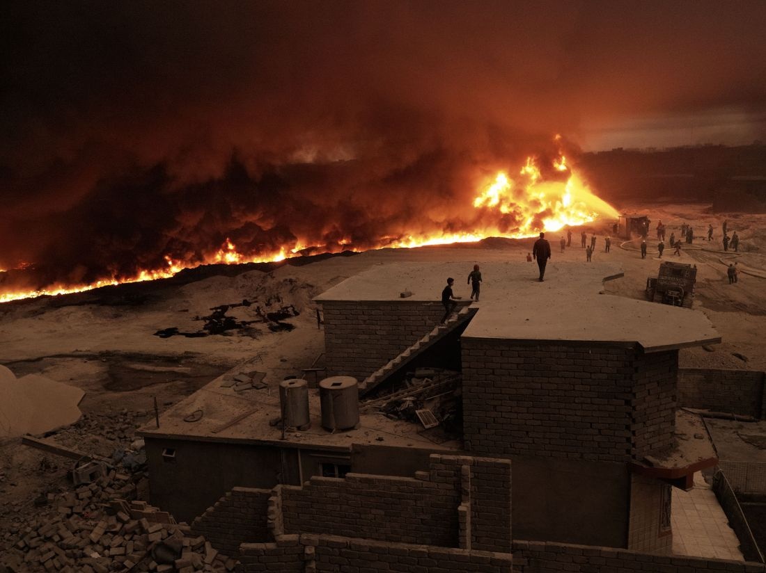 A family watches from their rooftop as firefighters struggle to extinguish a wall of flames creeping closer to their home. Qayyarah, Nineveh Governorate, Iraq, October 26, 2016. From [We Came From Fire?](?https://amzn.to/2L9l8Vm) by Joey L. – published by powerHouse Books